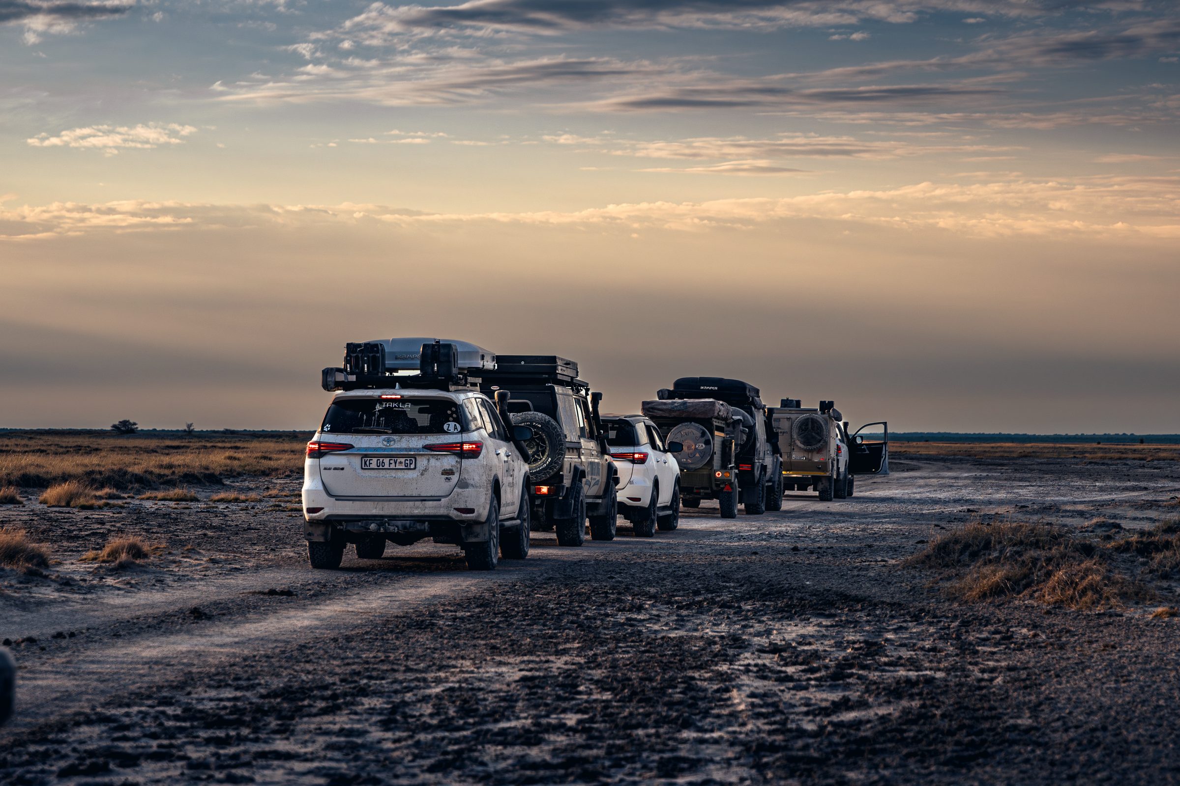 Five loaded 4x4s in convoy on a track across an open pan at sunset.