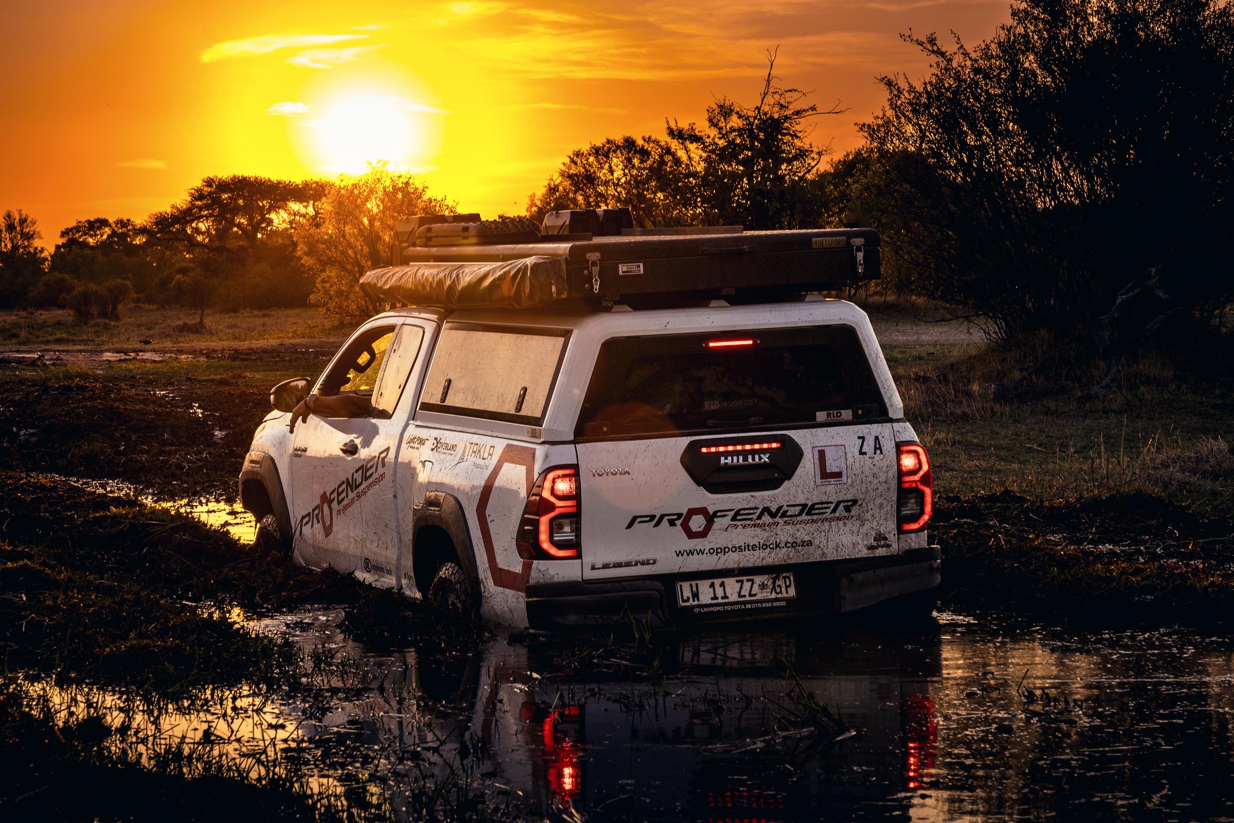 A double-cab bakkie loaded with a canopy, rooftop tent and roof boxes, standing in shallow water at sunset.