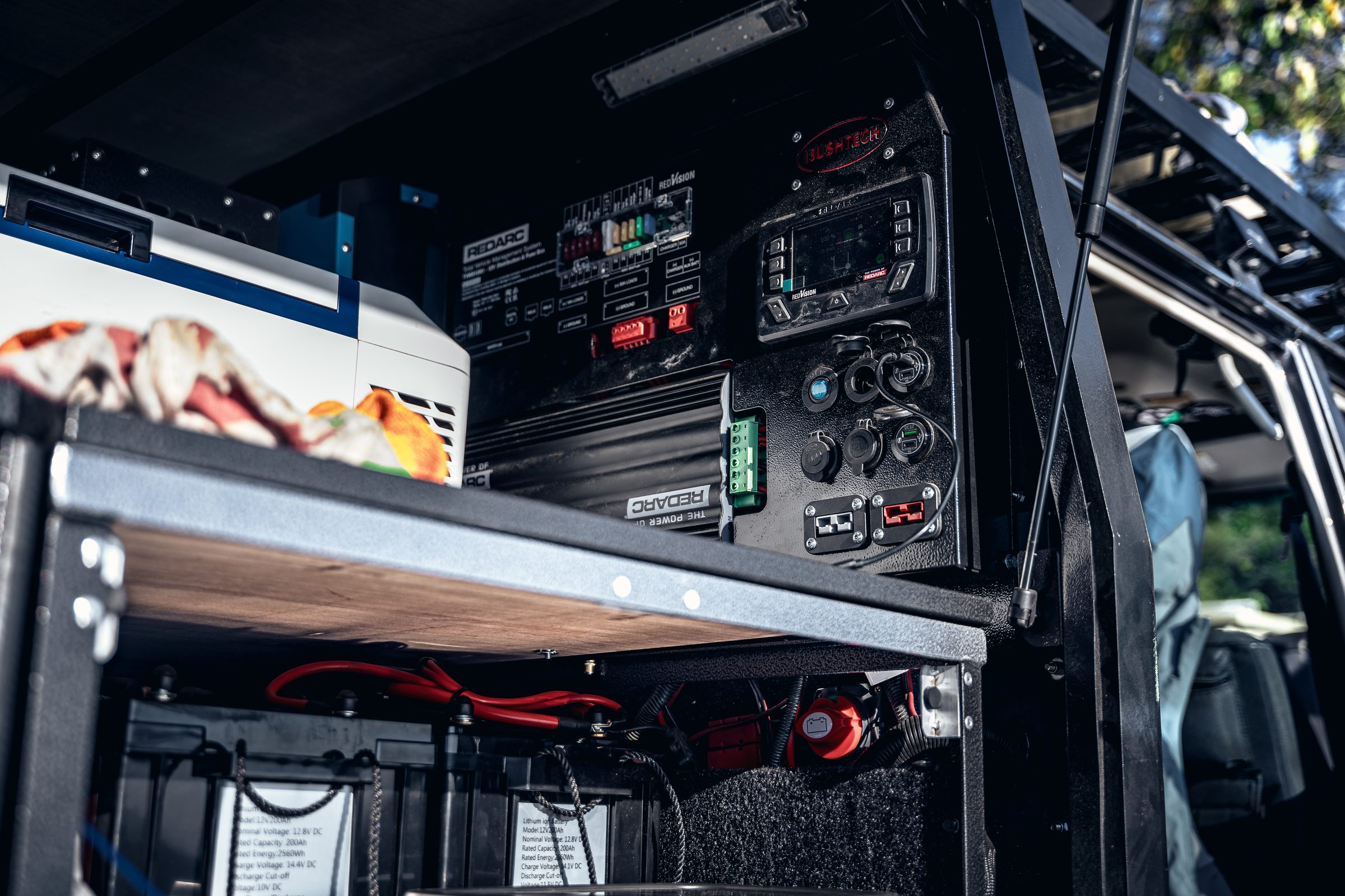 The inside of a vehicle canopy showing a 12V fridge on a slide, a control panel with switches and gauges, and two auxiliary batteries wired below.