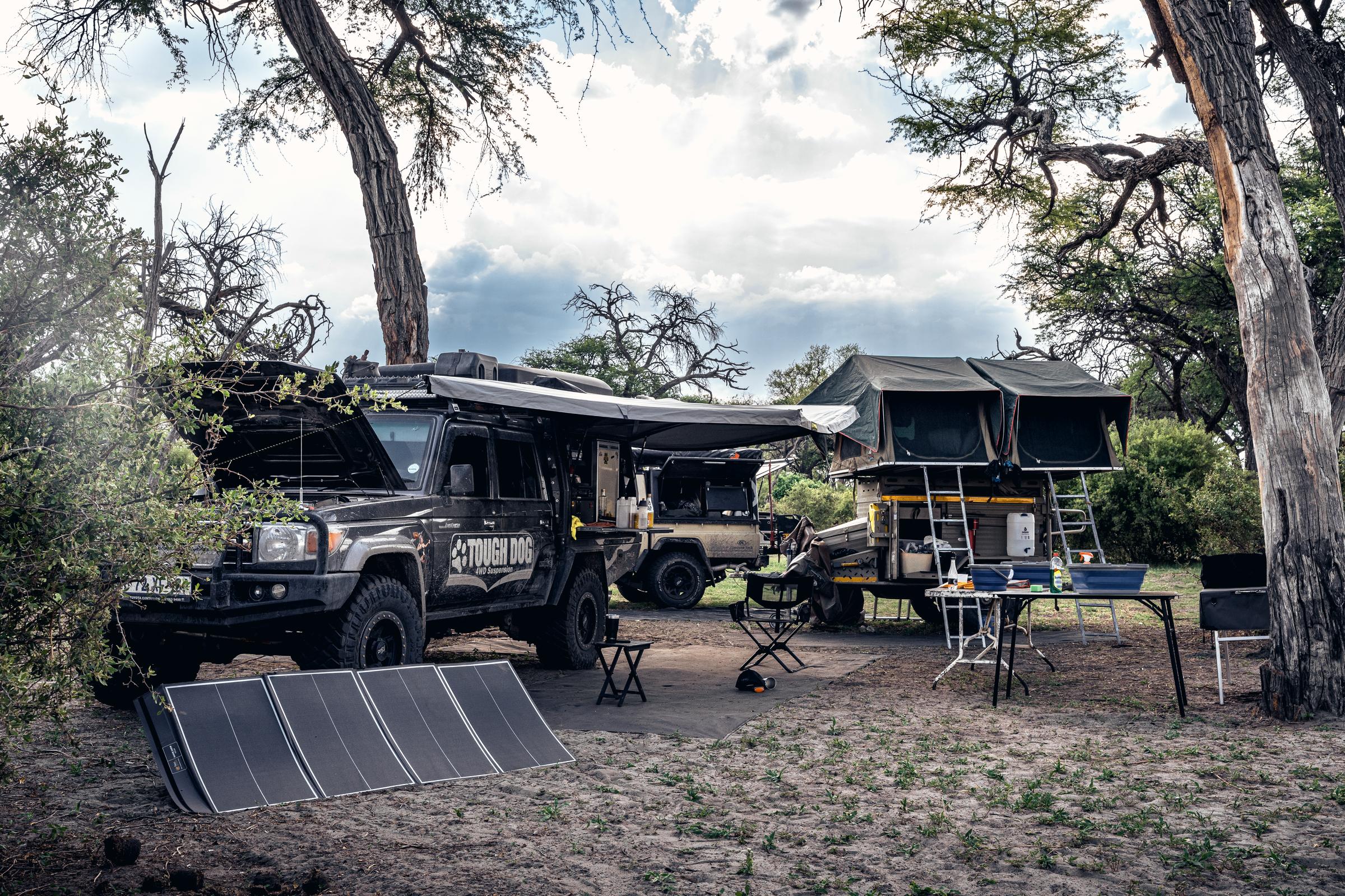 A bush camp under trees with two rooftop tents pitched, an awning out, camp chairs and a folding solar panel on the ground.